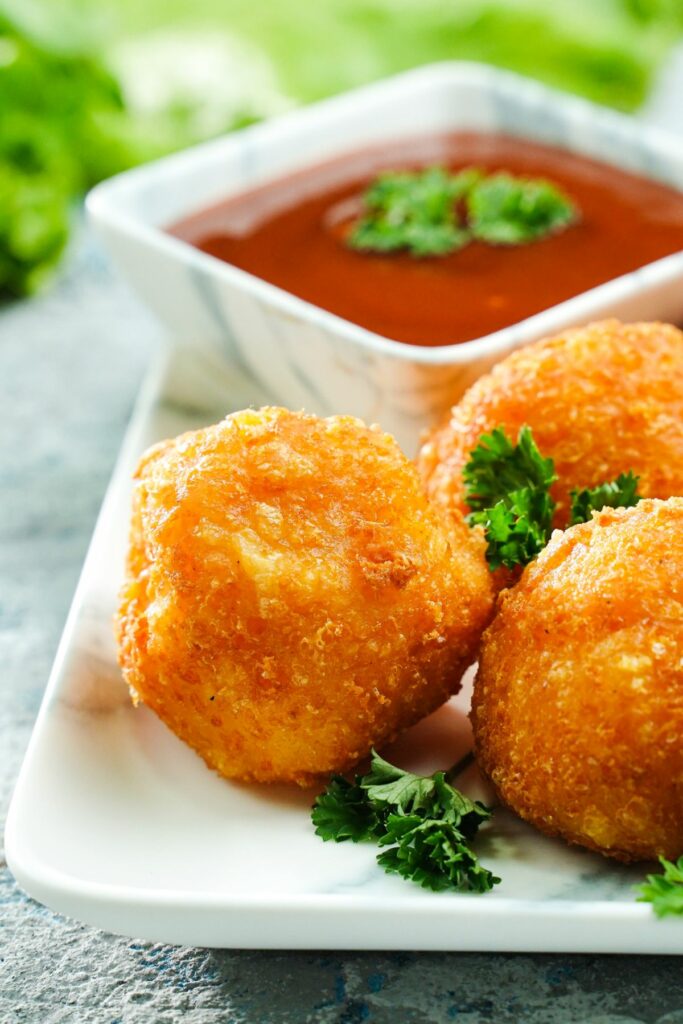 three cheese balls on corner of white plate with parsley and bowl of ketchup in background