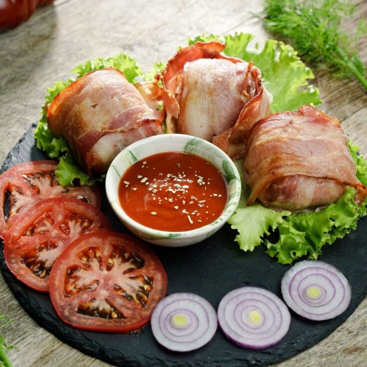 three bacon wrapped meatballs on lettuce next to tomato and onion on black plate on wood table