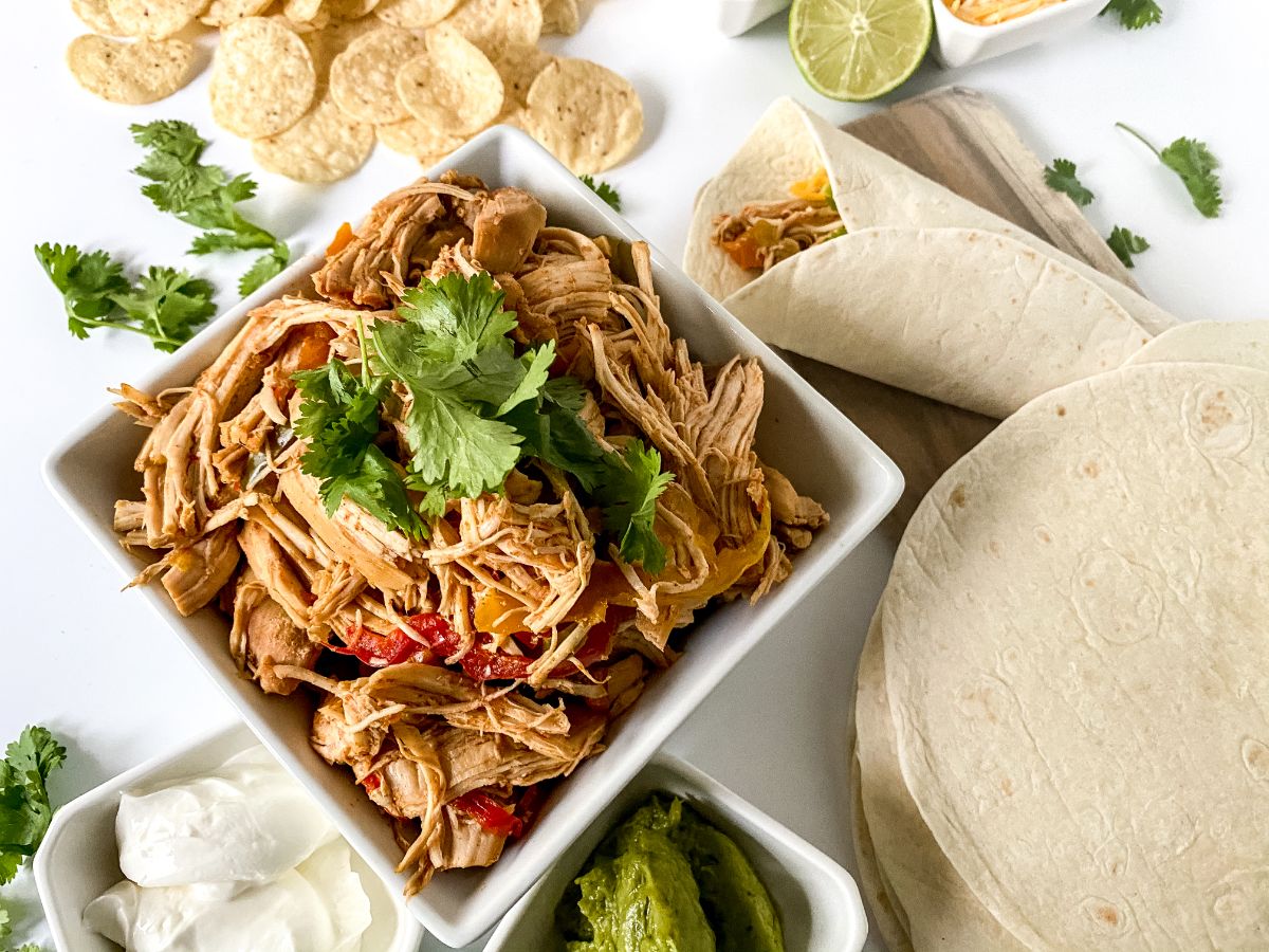 square white bowl of shredded chicken next to stack of tortillas on white table