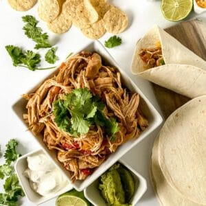 square white bowl of shredded chicken on white table with smaller bowls of sour cream and guacamole