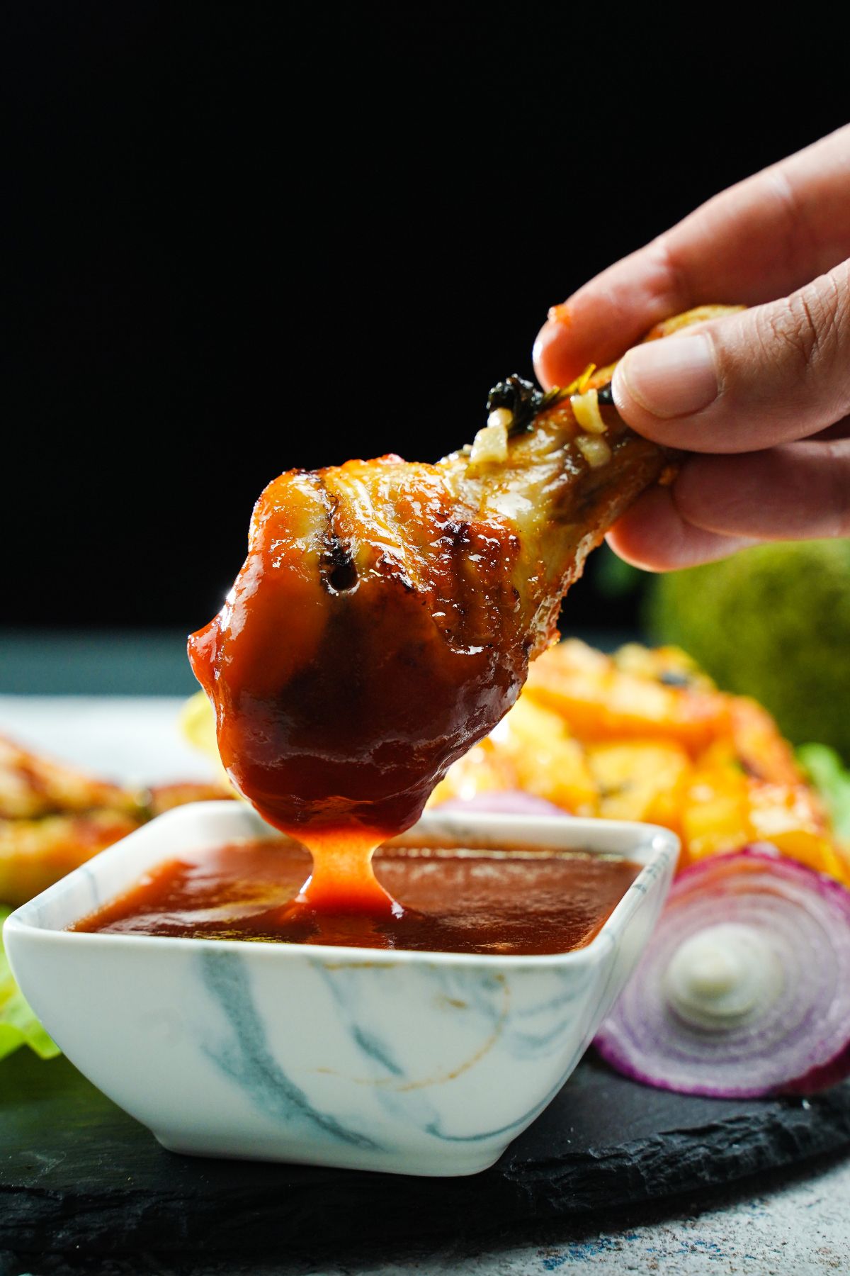 hand dipping chicken leg into sauce by black plate