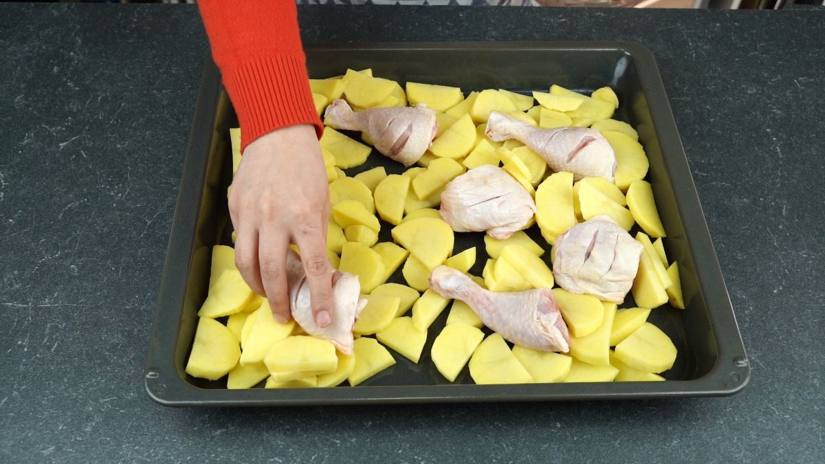 hand laying raw chicken legs onto sheet pan with potatoes
