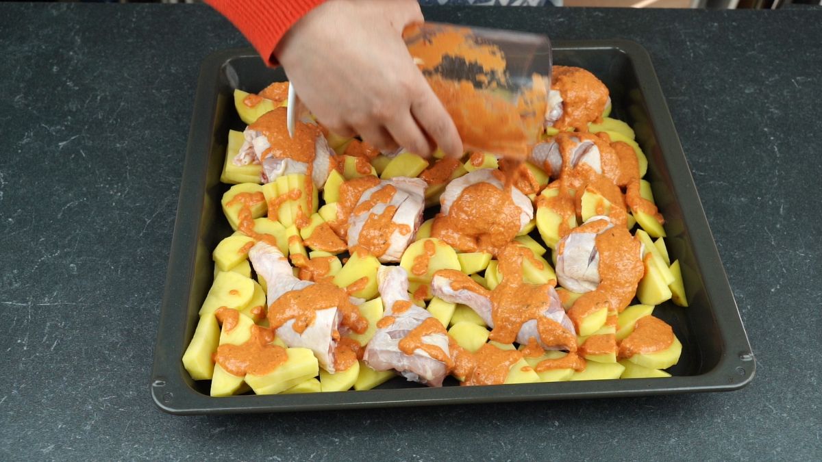hand pouring sauce over raw chicken legs on top of sliced potatoes on sheet pan