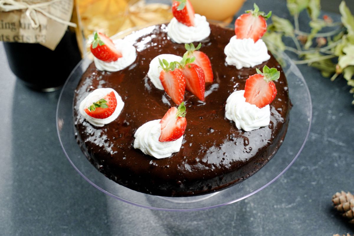 glass cake stand on gray table holding chocolate cake topped with whipped cream and strawberries
