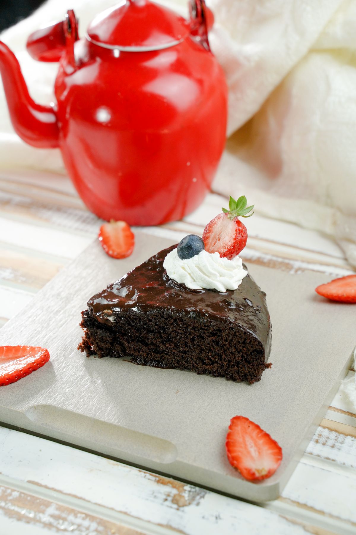 gray square plate holding single slice of chocolate cake with berries in front of red teapot