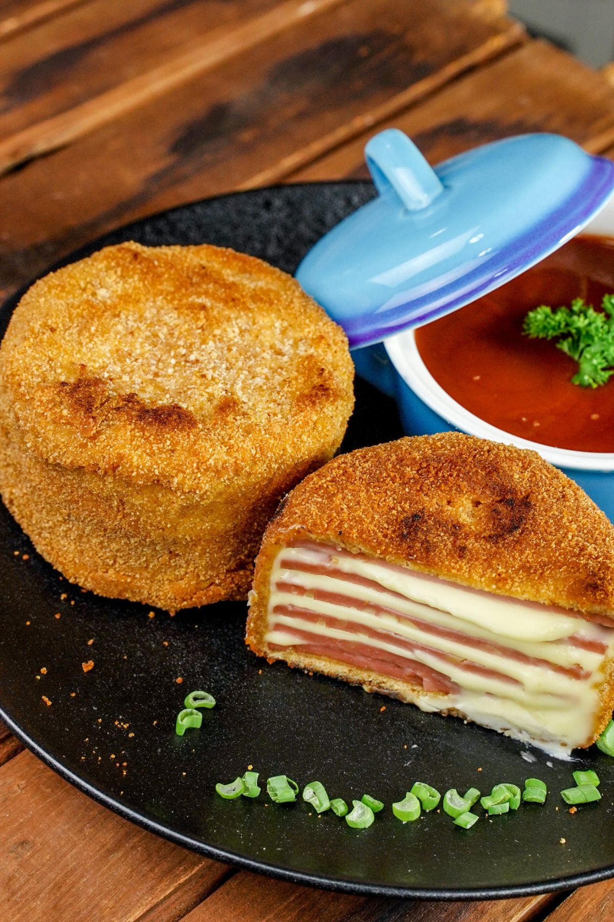 two ham cutlets on black plate with one cut in half and a blue bowl of soup on edge of plate