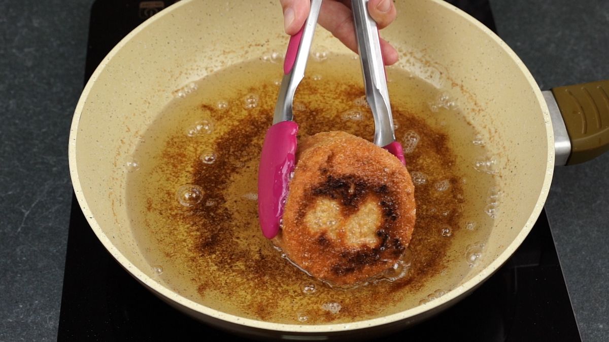 pink tongs turning ham and cheese cutlet in frying pan