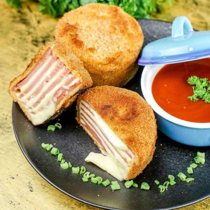 ham and cheese cutlet cut in half on black plate with bowl of tomato soup in background