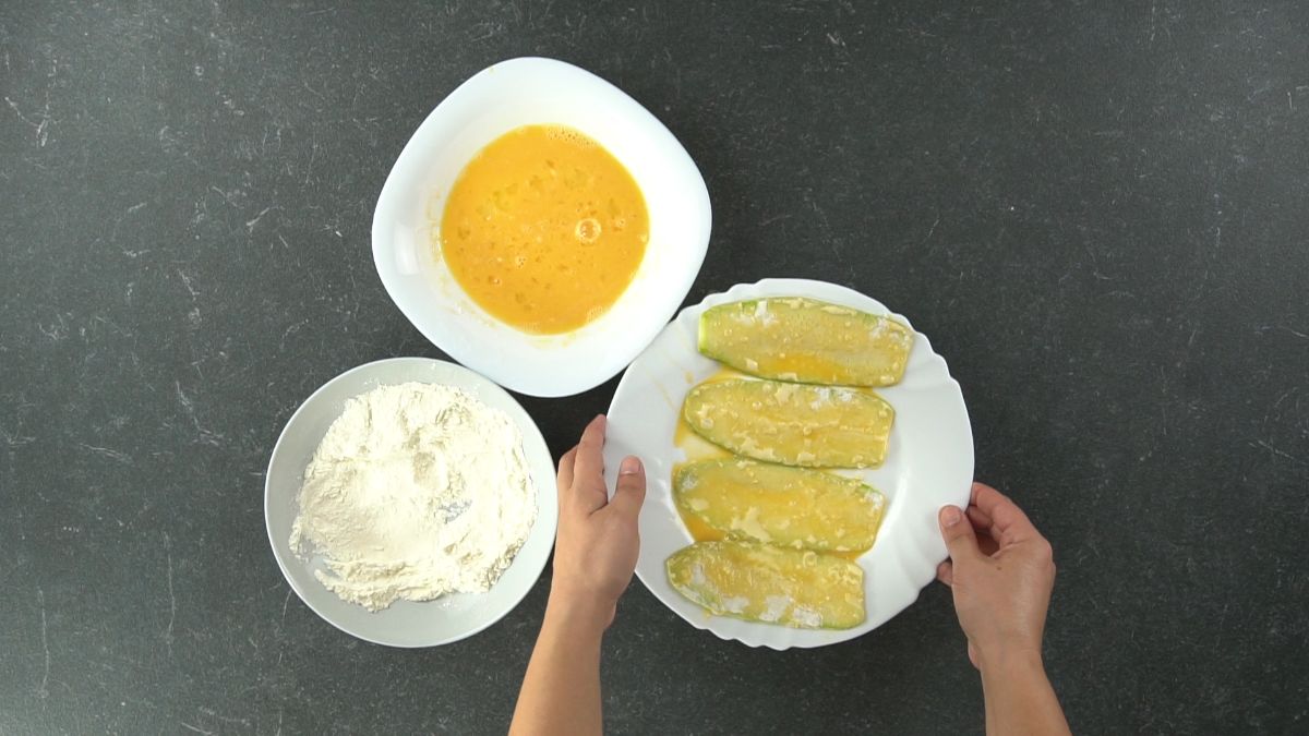 zucchini dipped in egg on white round plate next to bowl of flour and bowl of egg