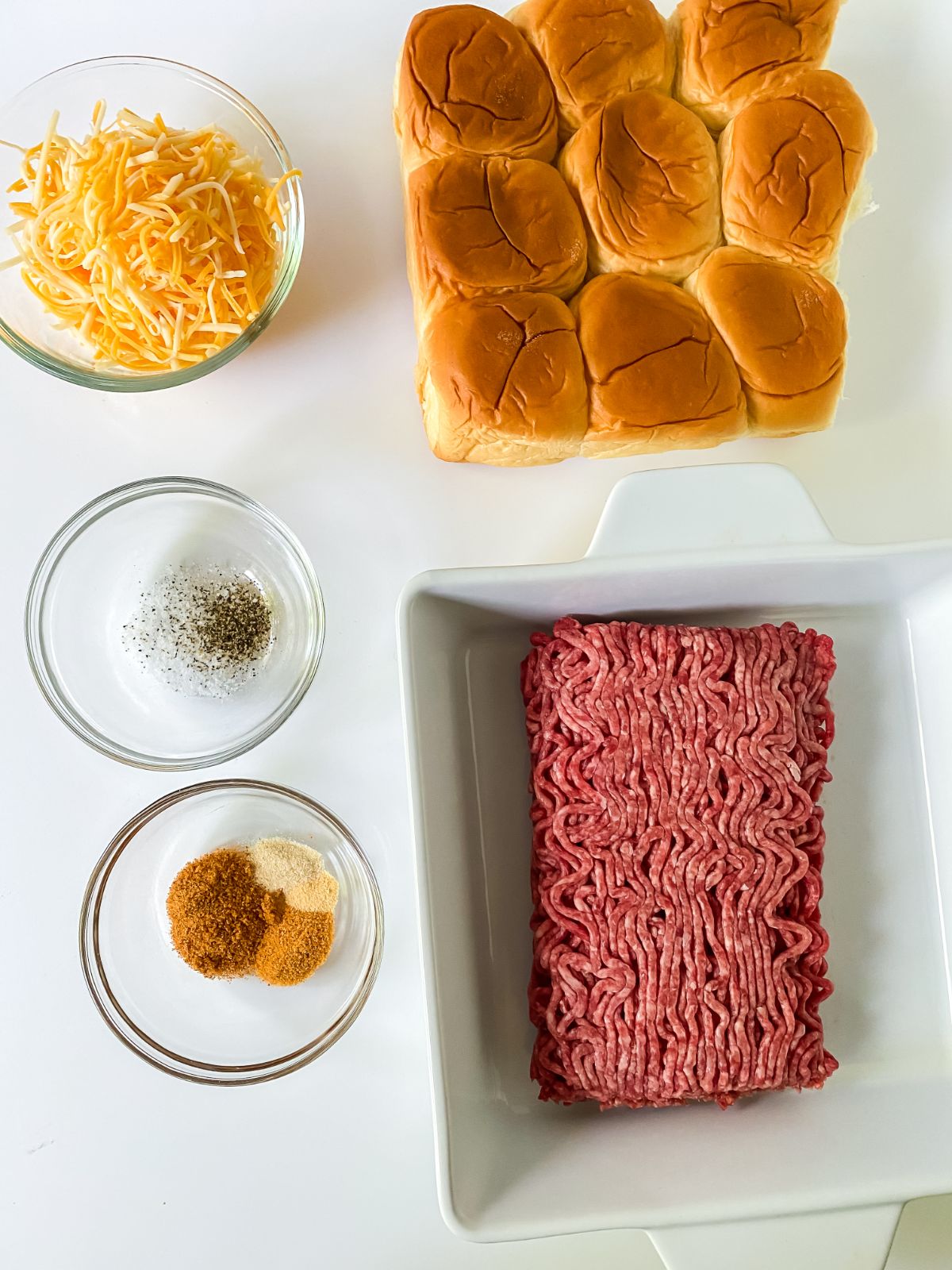 raw hamburger in dish next to seasonings in glass bowls and slider rolls on white table