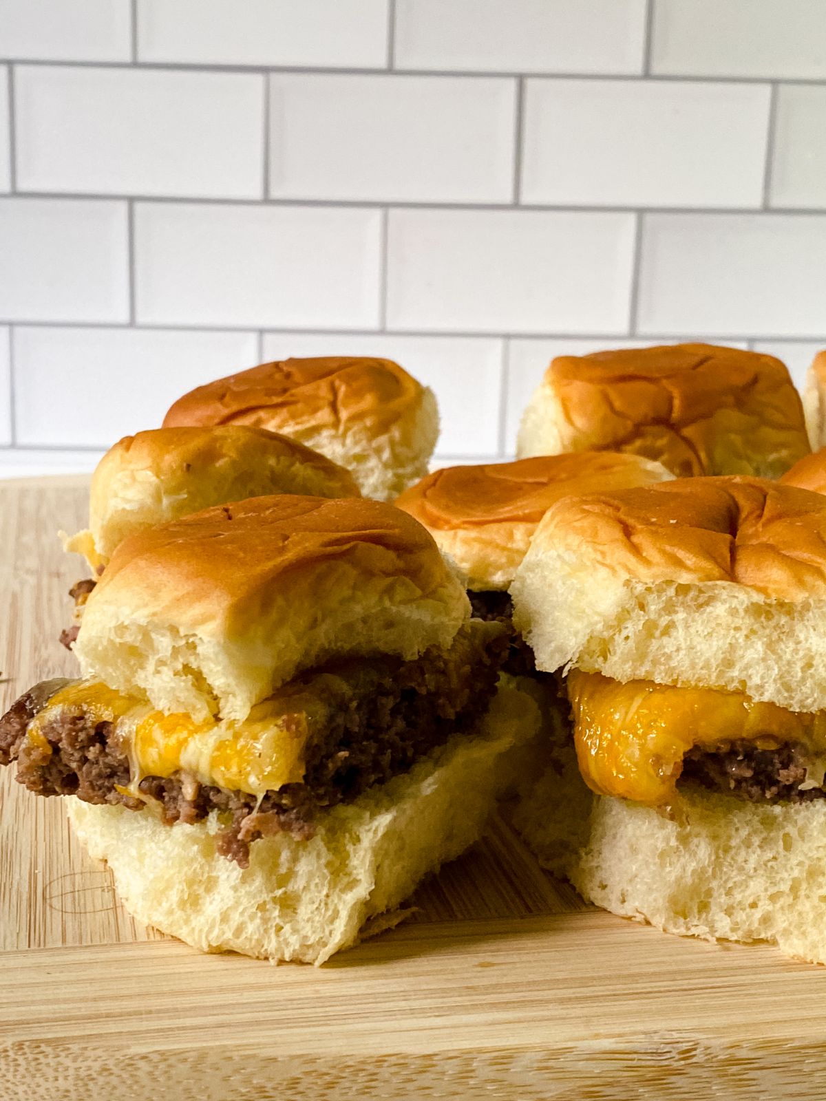 five cheeseburger sliders on cutting board in front of white subway tile