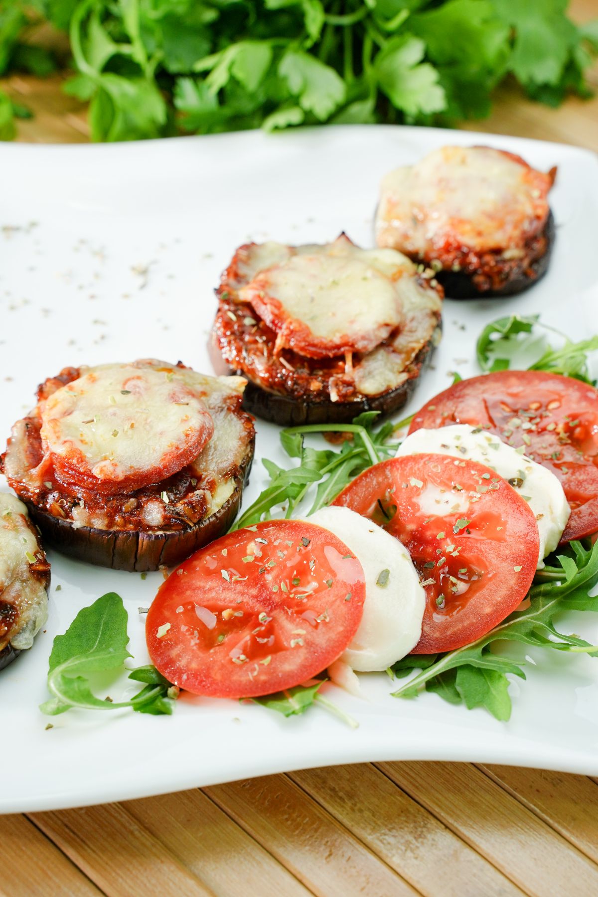 white plate of eggplant pizza bites with sliced tomato and cheese on arugula