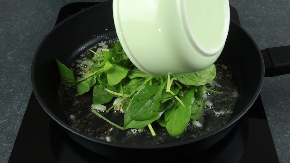 white bowl of spinach being dumped into black skillet