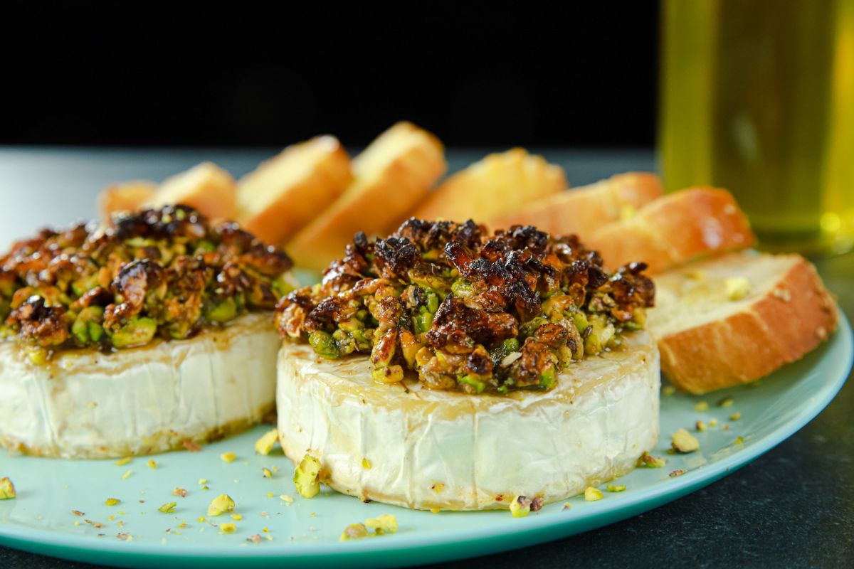 light teal plate with sliced baguettes and two rounds of brie topped with pistachios and honey