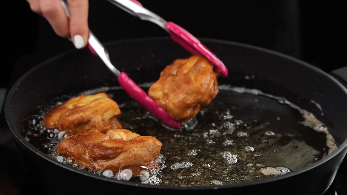pink tongs holding fried apple fritter above oil in black pan