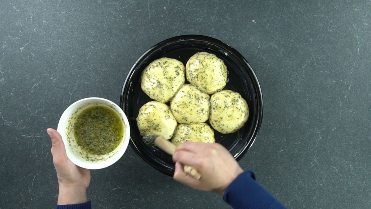 hand in blue shirt brushing herb butter over bread dough