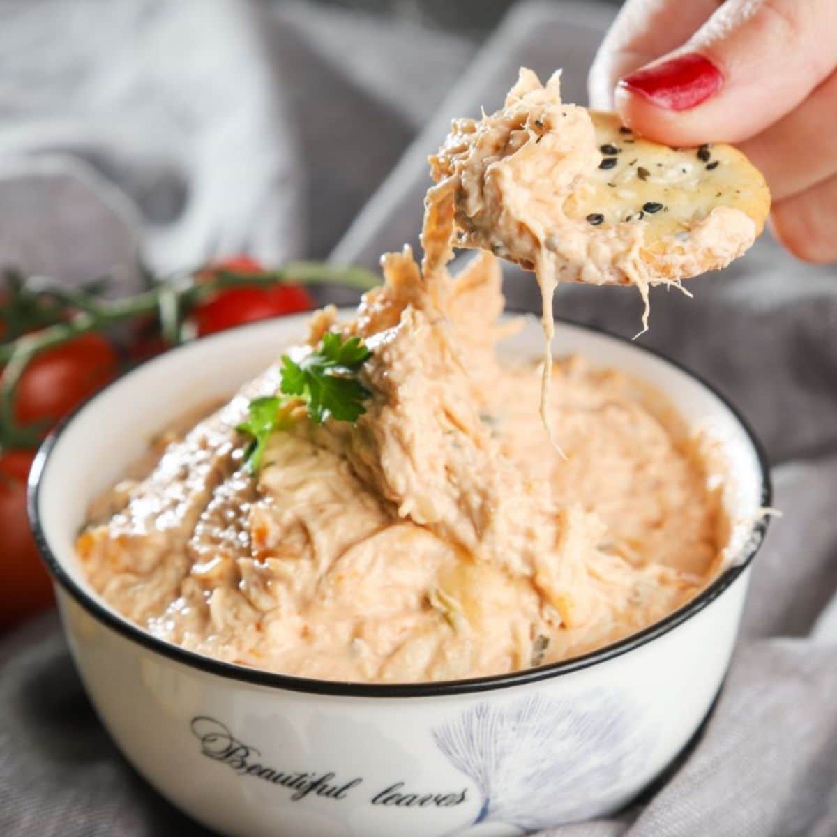 hand with red nails dipping into white bowl of dip with cracker