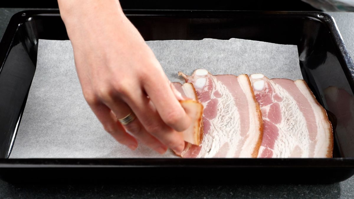 hand placing uncooked bacon onto baking sheet