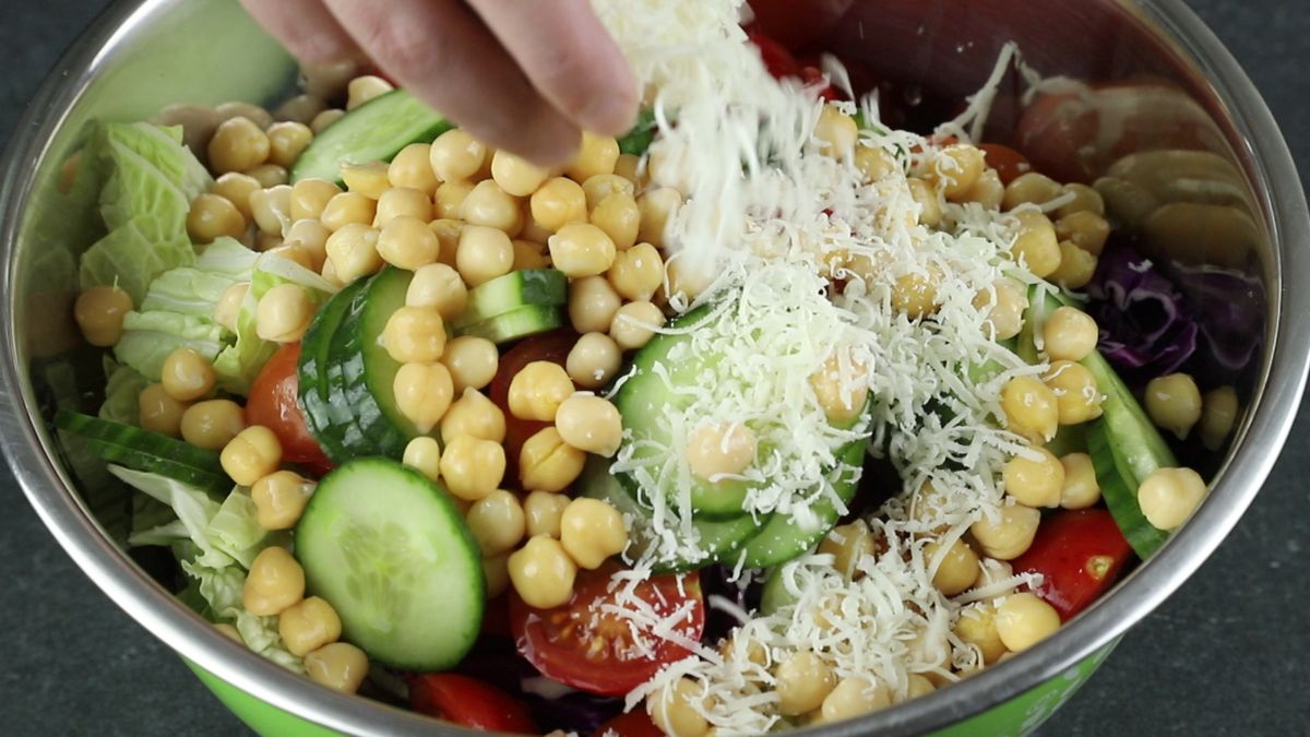 hand adding shredded cheese to bowl of chickpeas cucumber and tomato