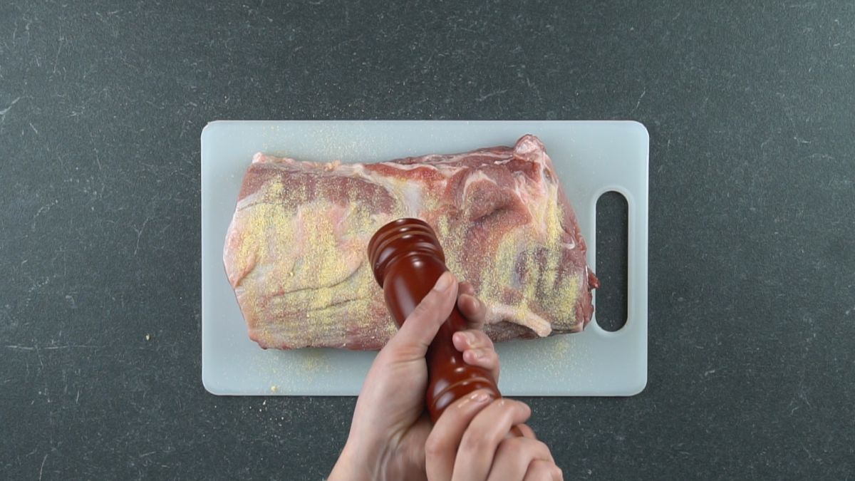 hand holding pepper mill above pork loin on white cutting board