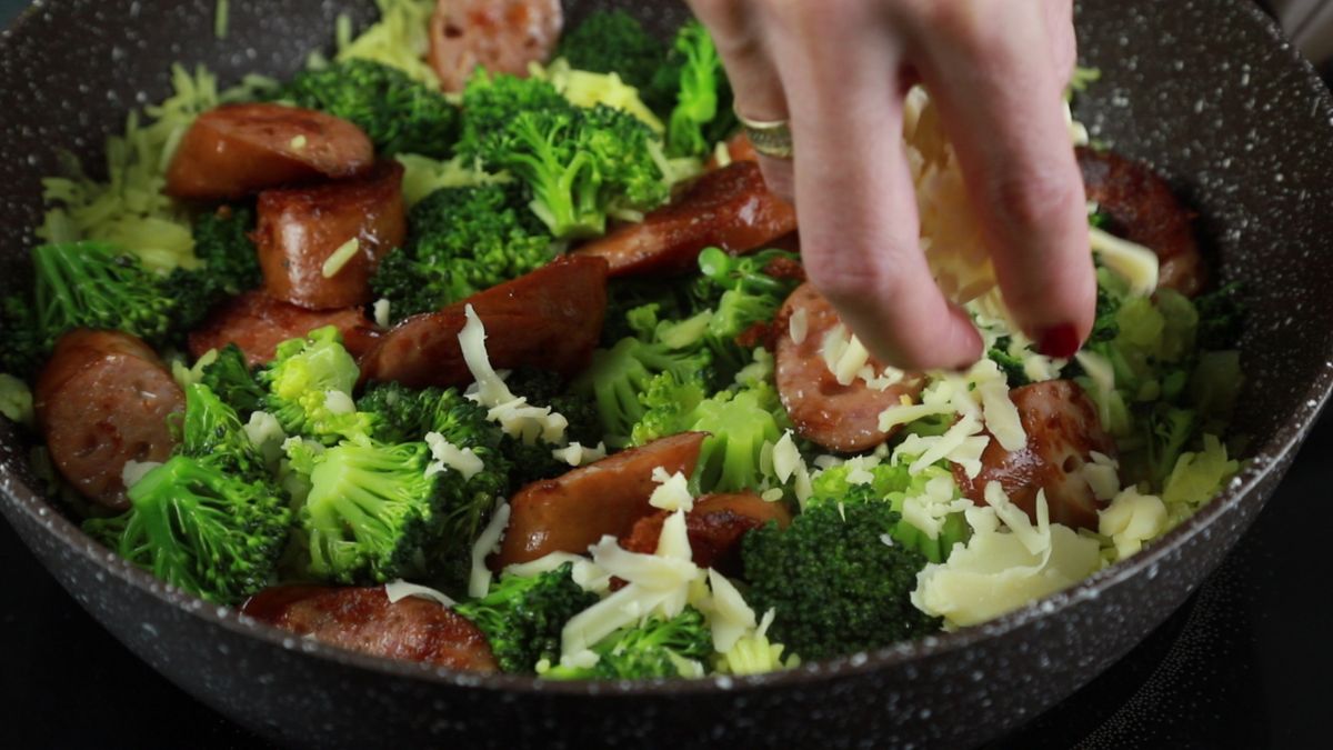 hand adding sausage and broccoli to skillet of rice