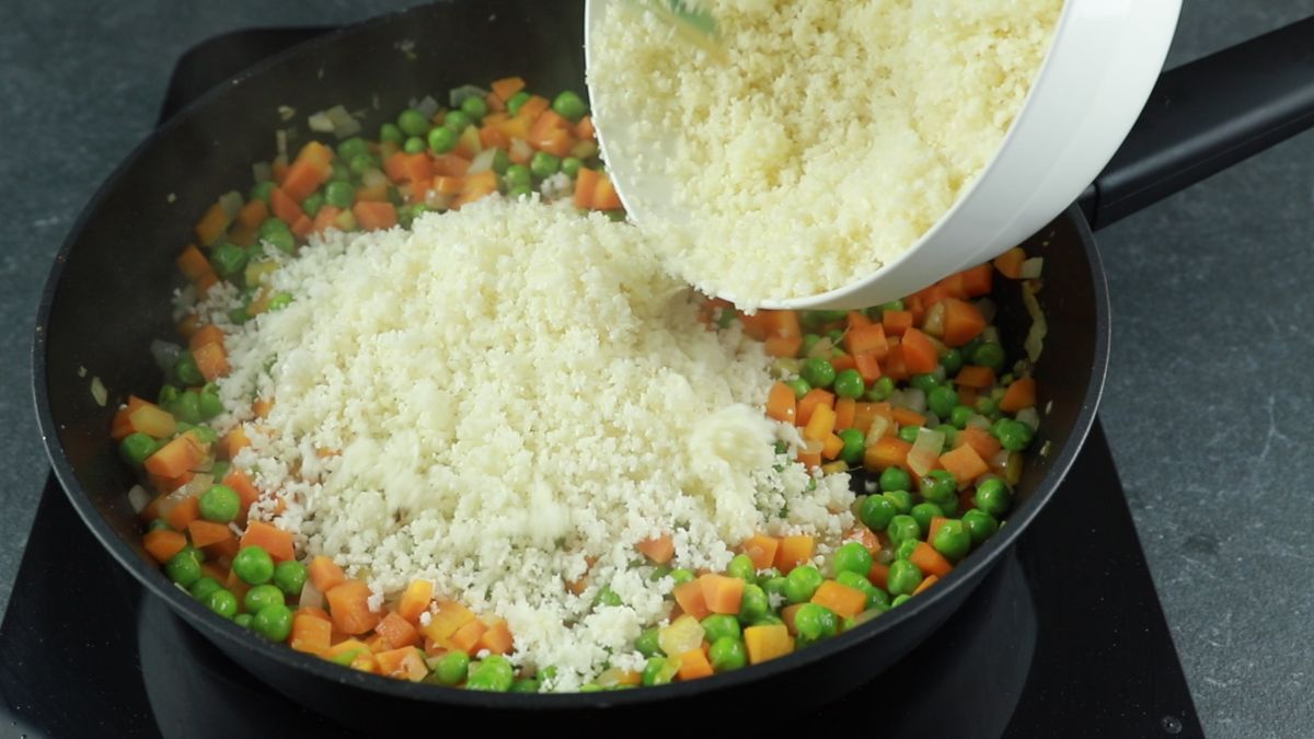 riced cauliflower being poured into black skillet with peas and carrots