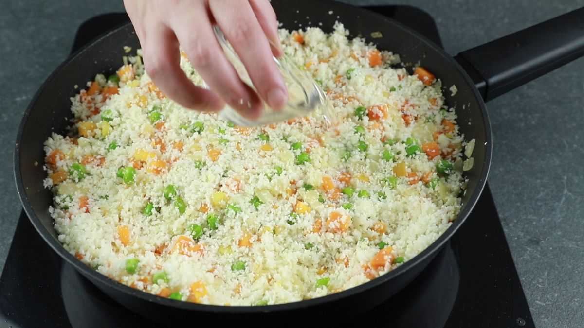 hand pouring salt into skillet of riced cauliflower