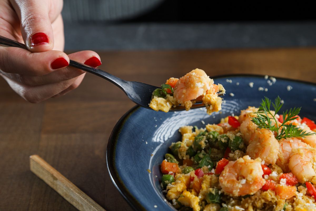 blue bowl of shrimp cauliflower fried rice on wood table with fork held above bowl