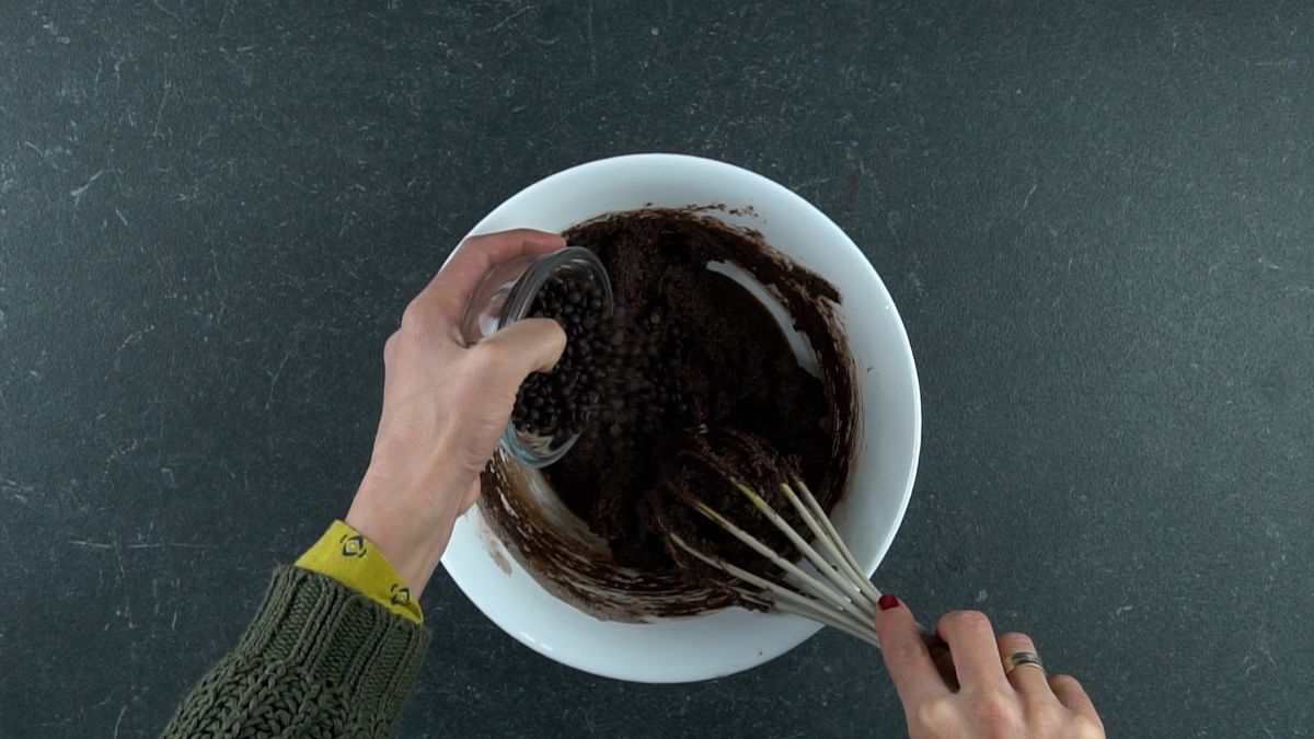 hand pouring chocolate chips into white bowl of brownie batter on gray table