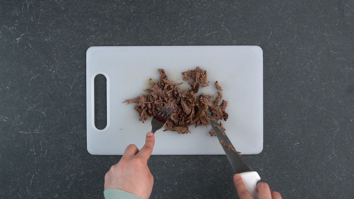hand holding knife and fork over beef on white cutting board