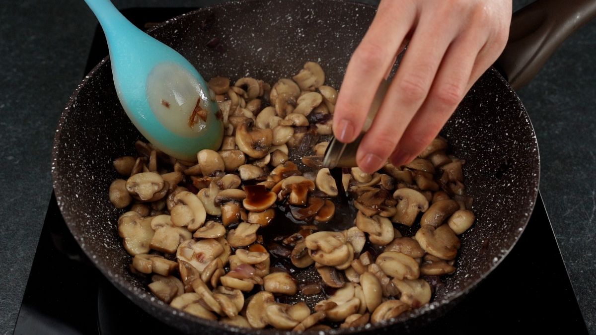 hand pouring sauce into skillet with cooked mushrooms and blue spoon