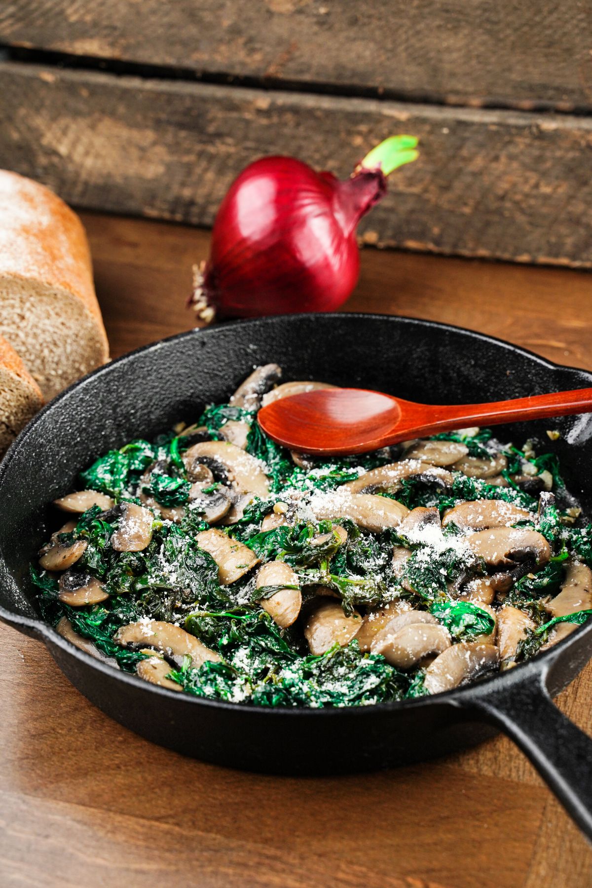cast iron skillet of mushrooms and spinach on wood table with red wooden spoon on corner of pan