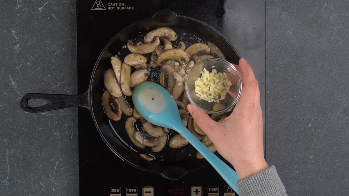 garlic being added to skillet of mushrooms with light teal spoon