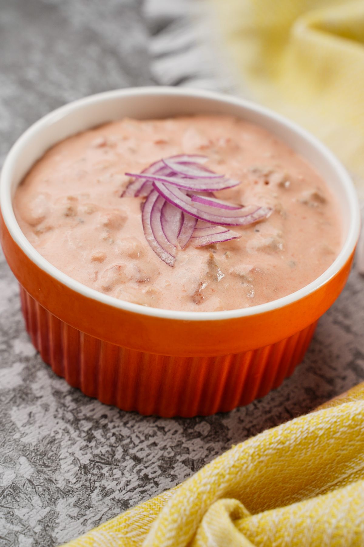 orange and white bowl of sausage dip with onion on top sitting on gray table