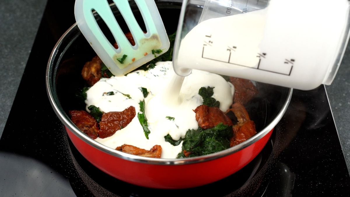glass cup of cream being ppoured into skillet with spinach and tomatoes