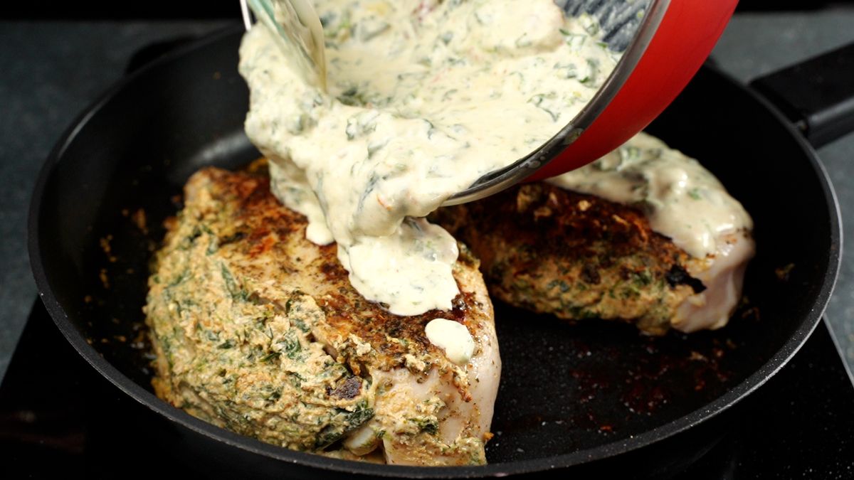 red pan of cream sauce being poured over stuffed chicken in black skillet