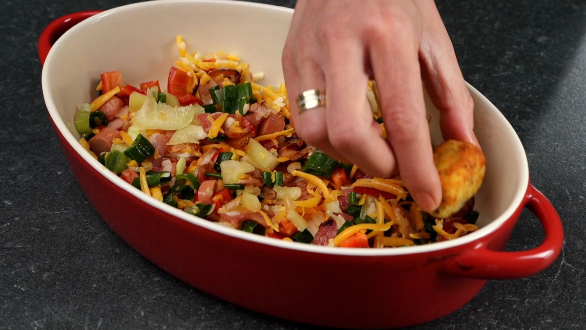 hand placing tater tots on top of vegetables in red oval dish