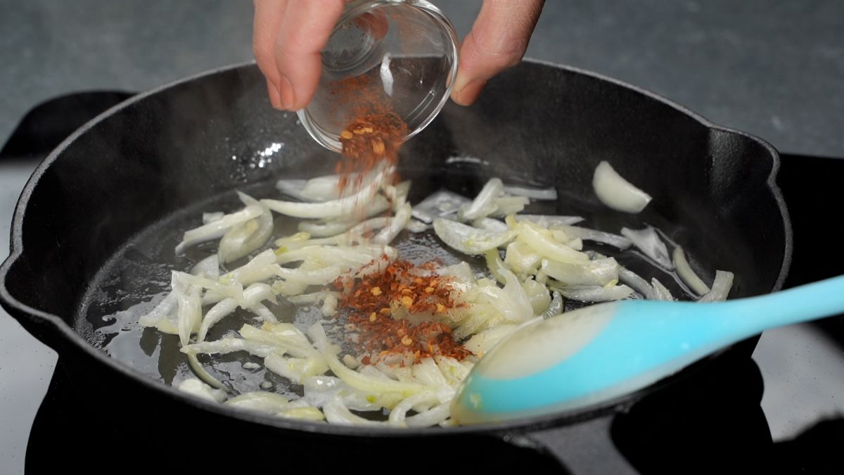 red pepper flakes being poured into skillet with onions