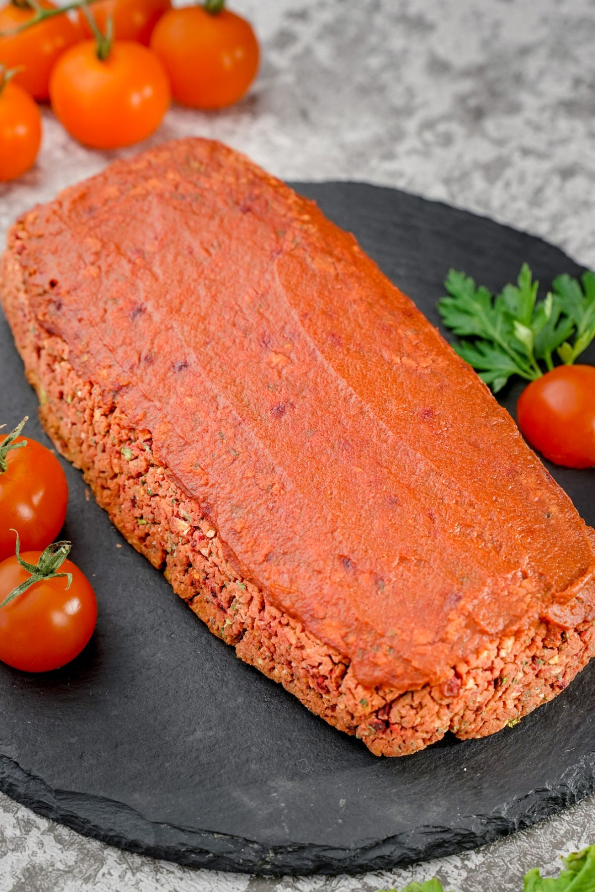 black round slate holding whole meatloaf with tomato ketchup on top and tomatoes on side