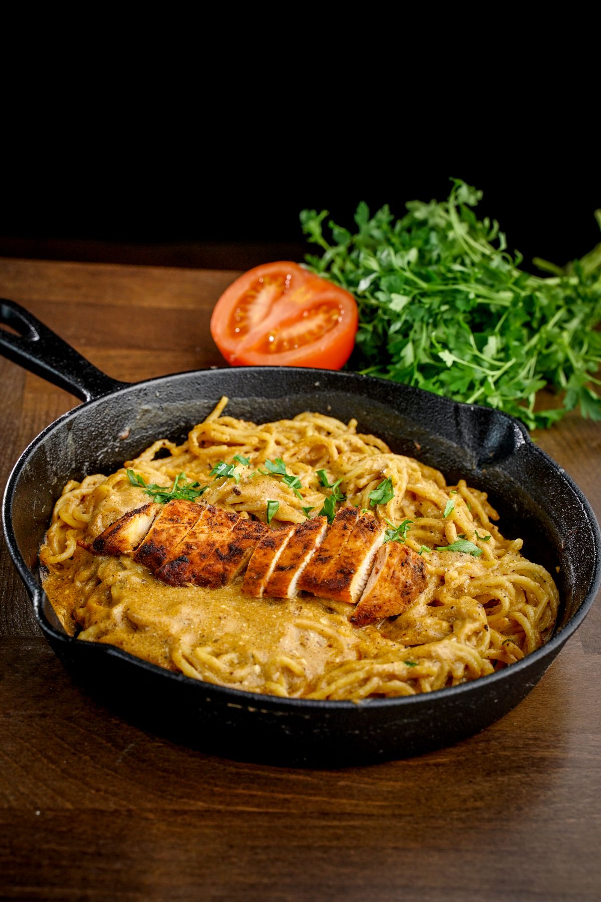 cast iron skillet of pasta with sliced chicken on wood table with black background