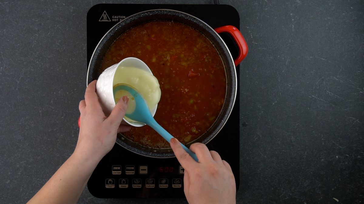 blue spoon scooping soup out of white bowl into tomato skillet