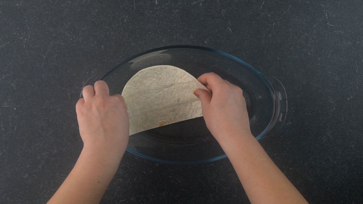 hand putting tortilla in bottom of glass casserole dish