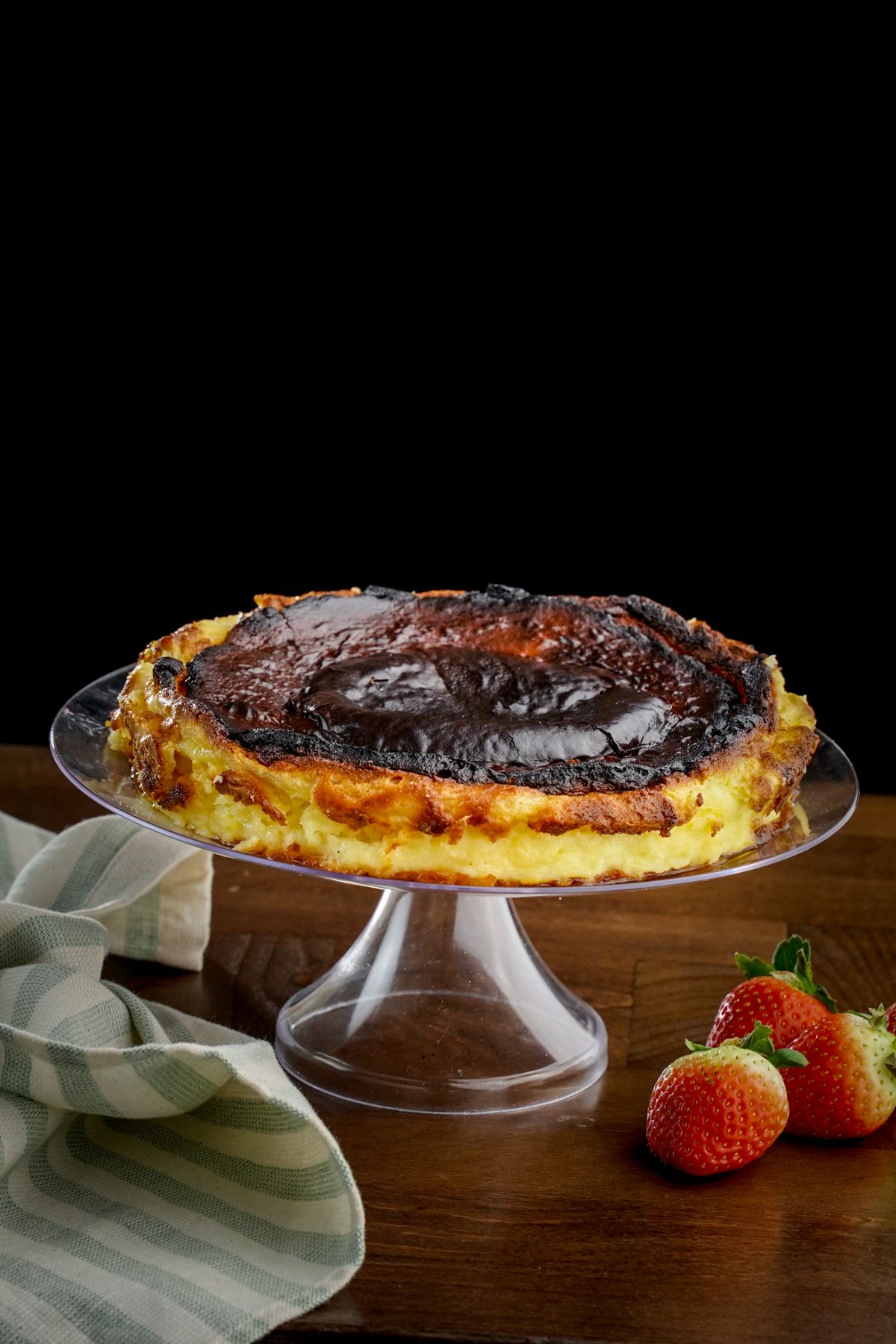 cake stand holding burnt basque cheesecake with black background on gray napkin with strawberries on table