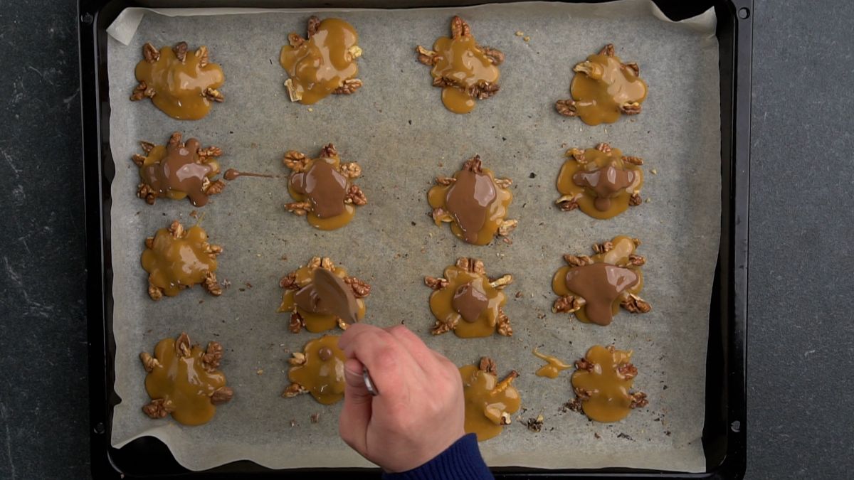 hand spooning chocolate over caramel turtles on baking sheet