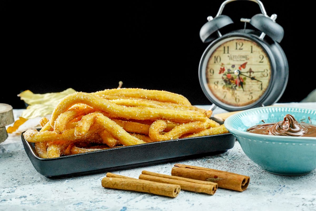 churros stacked in black tray next to blue bowl of caramel sauce with black background