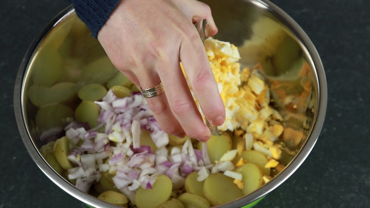hand pouring red onion and egg into pot with potatoes