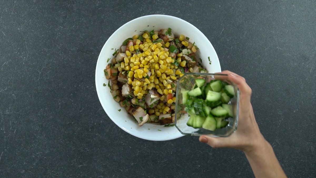 cucumber and corn being added to bowl of chicken chickpea salad