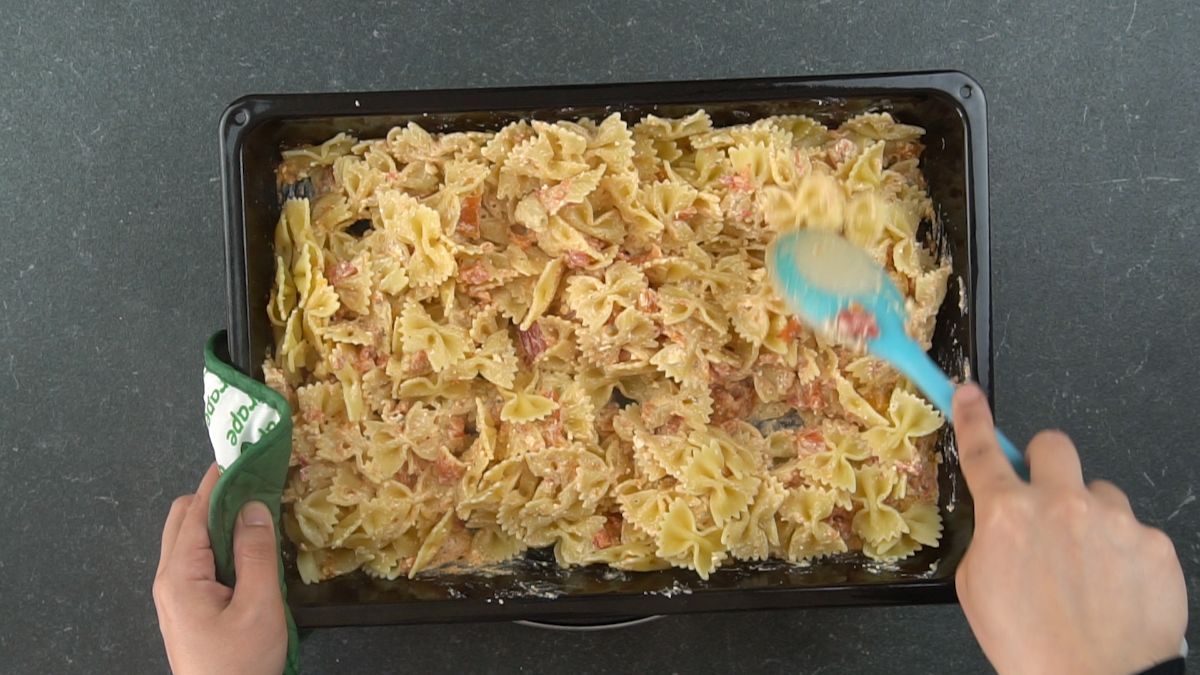 blue spoon stirring pasta into baked tomato and feta sauce