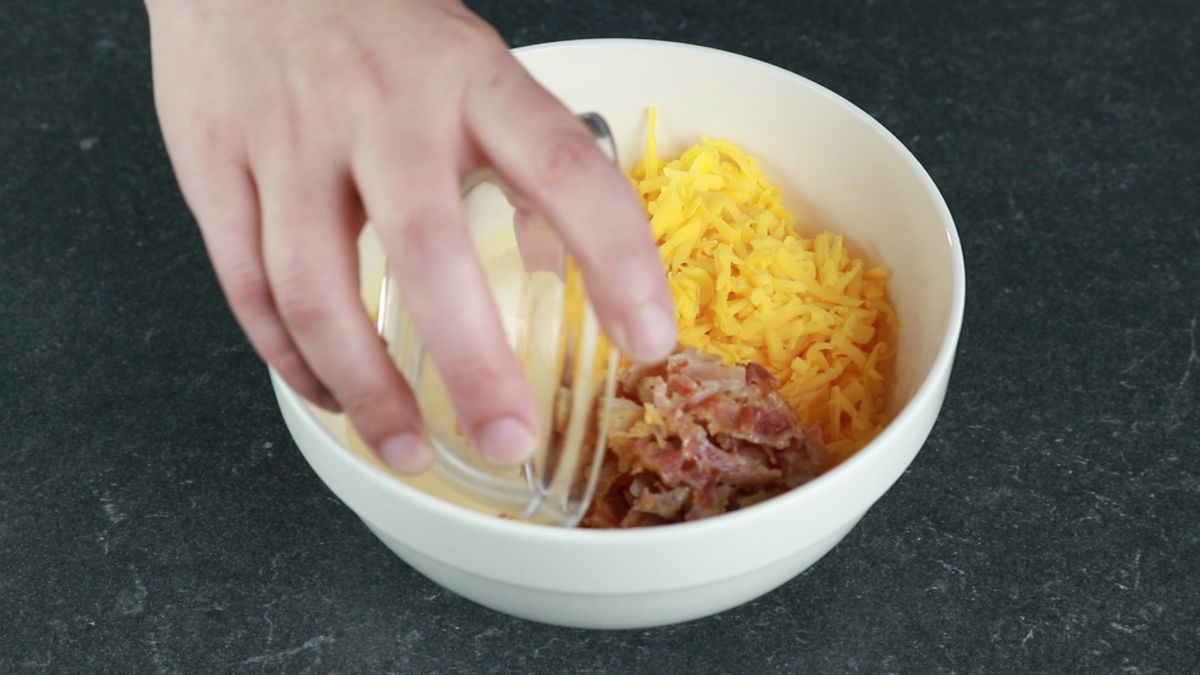 bacon being added to white bowl with eggs and cheese