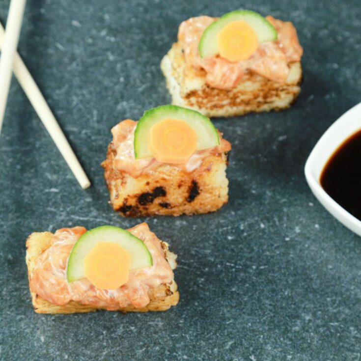 three pieces crispy rice with salmon topped by cucumber on gray table